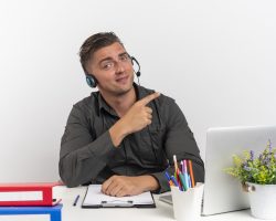 young confident blonde office worker man on headphones sits at desk with office tools using laptop points at side isolated on white background with copy space